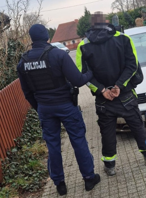 na chodniku przed osobowym seatem stoją policjant i zatrzymany mężczyzna. Mężczyzna ma ręce skute za plecami kajdankami. Policjant trzyma go za ramię