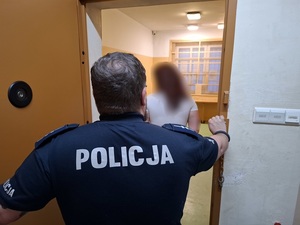 w policyjnej celi stoi kobieta, skierowana tyłem do obiektywu. Za nią przy otwartych drzwiach celi stoi policjant służby dyżurnej