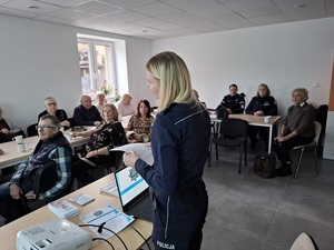 w sali przy stole siedzą seniorzy, pracownica socjalna i dwoje policjantów. Przed nimi i przy laptopie stoi policjantka