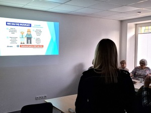 w sali przy stole siedzą seniorzy. Na ścianie wyświetla się slajd "nie daj się oszukać". Obok stoi policjantka