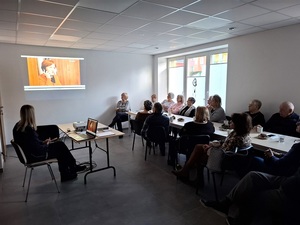 w sali przy stole siedzą seniorzy. Na ścianie wyświetla się film. Naprzeciw seniorów siedzi policjantka