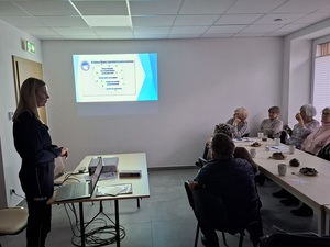 w sali przy stole siedzą seniorzy. Na ścianie wyświetla się slajd z Krajową Mapą Zagrożeń Bezpieczeństwa. Naprzeciw seniorów stoi policjantka