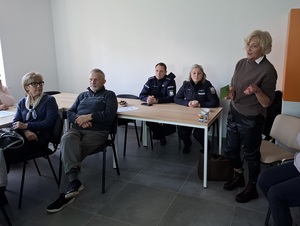 w sali przy stole siedzą seniorzy oraz dwoje policjant i policjantka. Obok stoi pracownica socjalna, która przemawia do zgromadzonych