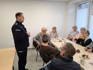 w sali przy stole siedzą seniorzy. Obok stoi policjant, który przemawia do seniorów