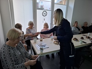 w sali przy stole siedzą seniorzy. Policjantka rozdaje im ulotki