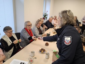 w sali przy stole siedzą seniorzy. Dzielnicowa rozdaje im elementy odblaskowe