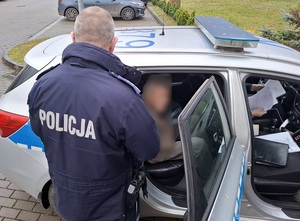 na tylnym siedzeniu radiowozu siedzi zatrzymany mężczyzna. Przed nim, przy otwartych drzwiach radiowozu stoi policjant