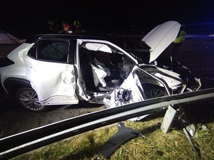 Pora wieczorowa. Na poboczu, przy uszkodzonej barierce znajduje się biała osobowa toyota z uszkodzeniami powypadkowymi. Karoseria z boku pojazdu jest zgnieciona. Na drodze leżą drobne fragmenty auta