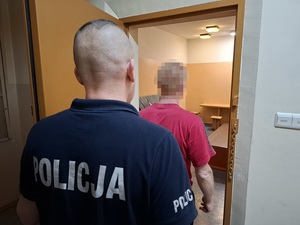 w celi stoi mężczyzna zwrócony tyłem do obiektywu. Za nim przy otwartych drzwiach celi stoi policjant
