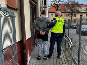 przed drzwiami budynku komendy stoi policjantka, trzymająca za ramię mężczyznę, który ma ręce skute za plecami kajdankami