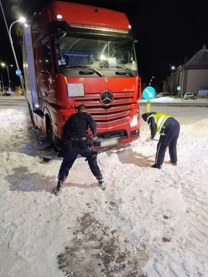 na dworze ciemno. Na zaśnieżonej jezdni stoi ciężarówka z naczepą. Dotyka przechylonej lampy. Policjant i policjantka łopatami odgarniają śnieg spod kół ciężarówki