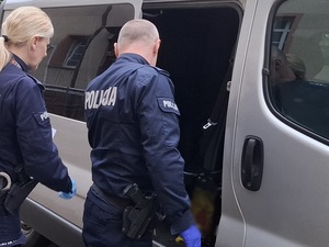 dwoje policjantów służby konwojowej stoi przed nieoznakowanym radiowozem. Policjant trzyma otwarte boczne drzwi radiowozu