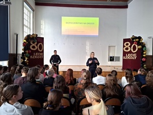 w szkolnej auli przed siedzącymi w rzędach uczniami stoją policjant ruchu drogowego i policjantka. Za nimi na ekranie wyświetla się napis "Bezpieczeństwo na drodze"