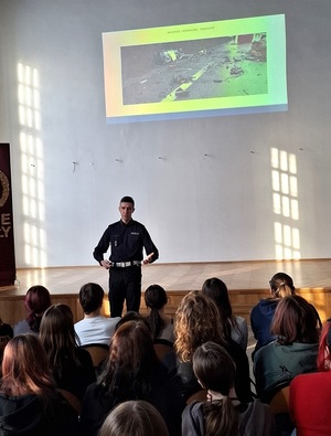 w szkolnej auli przed siedzącymi w rzędach uczniami stoi policjant ruchu drogowego. Za nimi na ekranie wyświetla się zdjęcie samochodu po wypadku