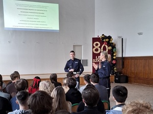 w szkolnej auli przed siedzącymi w rzędach uczniami stoją policjant ruchu drogowego i policjantka. Za nimi na ekranie wyświetla się treść prezentacji