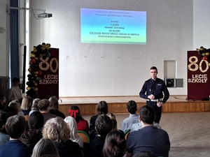w szkolnej auli przed siedzącymi w rzędach uczniami stoi policjant ruchu drogowego. Za nimi na ekranie wyświetla się prezentacja