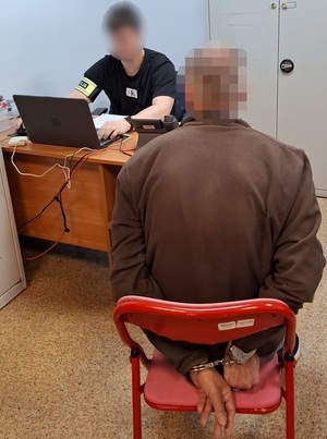 w policyjnym pokoju służbowym na krześle siedzi mężczyzna, który ma ręce skute za plecami kajdankami. Przed nim przy biurku, przed laptopem siedzi policjant służby kryminalnej.