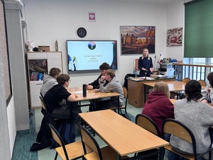 w klasie szkolnej przed siedzącymi w ławkach uczniami stoi policjantka. Za nią na ekranie wyświetla się prezentacja