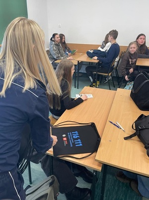 w klasie szkolnej policjantka rozdaje siedzącym w ławkach uczniom czarne worki szkolne z nadrukowanym numerem telefonu zaufania dla dzieci i młodzieży