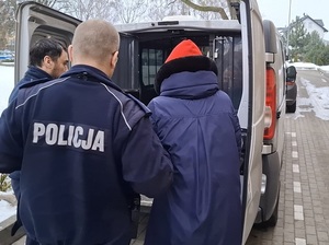 przed otwartymi okratowanymi drzwiami z tyłu nieoznakowanego radiowozu stoi zatrzymana kobieta. Policjant trzyma ją za ramię. Drugi policjant przytrzymuje drzwi
