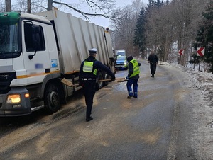 na oblodzonej jezdni, na poboczu stoi ciężarówka. Policjant i mężczyzna ubrany w odblaskowy strój roboczy przy użyciu łopat rozsypują piasek na jezdnię