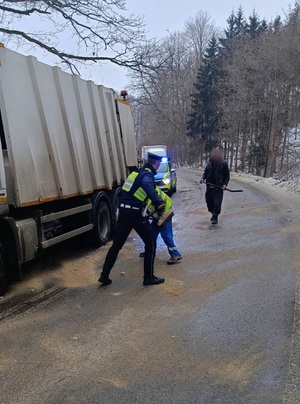 na oblodzonej jezdni, na poboczu stoi ciężarówka. Policjant i mężczyzna ubrany w odblaskowy strój roboczy przy użyciu łopat rozsypują piasek na jezdnię