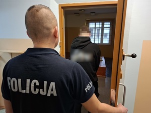w otwartych drzwiach celi stoi mężczyzna skierowany tyłem do obiektywu. Za nim stoi policjant służby dyżurnej, który przytrzymuje drzwi celi