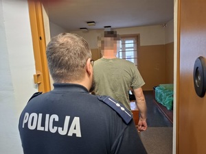 w otwartych drzwiach celi stoi mężczyzna. Za nim stoi policjant służby dyżurnej, który przytrzymuje drzwi