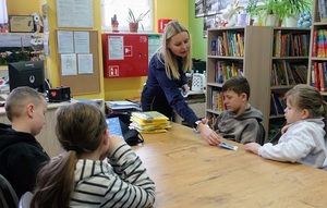 biblioteka. Policjantka rozdaje siedzącym przy stole dzieciom ulotki