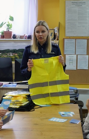 w bibliotece przy stole stoi policjantka, która prezentuje kamizelkę odblaskową