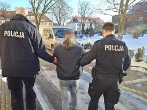 Zima. Na zewnątrz budynku komendy dwaj policjanci służby konwojowej prowadzą w kierunku nieoznakowanego radiowozu zatrzymanego mężczyznę