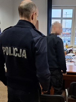 w pokoju przesłuchań stoi zatrzymany mężczyzna. Za nim stoi umundurowany policjant