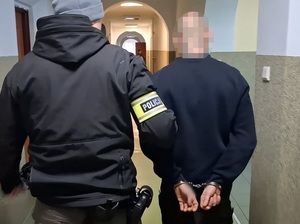 nieumundurowany policjant z opaską na ramieniu z napisem "Policja" prowadzi korytarzem mężczyznę, który ma ręce skute za plecami kajdankami