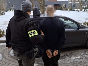 na zewnątrz budynku komendy nieumundurowany policjant z opaską na ramieniu z napisem "Policja" prowadzi w kierunku nieoznakowanego radiowozu mężczyznę, który ma ręce skute za plecami kajdankami