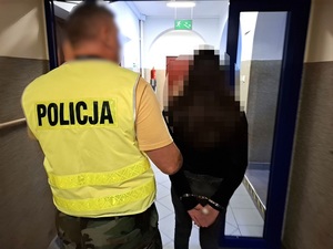 przez przeszklone drzwi przechodzą: kobieta, która ma ręce skute za plecami kajdankami oraz policjant trzymający ją za przedramię