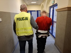 policjant w kamizelce odblaskowej z napisem "policja" prowadzi korytarzem mężczyznę, który ma ręce skute za plecami kajdankami