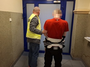 na korytarzu policjant w kamizelce odblaskowej z napisem "policja", przytrzymujące mężczyznę, który ma ręce skute za plecami kajdankami, otwiera drzwi prowadzące do pomieszczenia dla osób zatrzymanych
