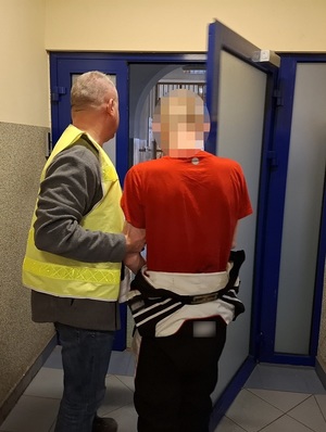 na korytarzu policjant w kamizelce odblaskowej z napisem "policja", przytrzymujące mężczyznę, który ma ręce skute za plecami kajdankami, otwiera drzwi prowadzące do pomieszczenia dla osób zatrzymanych