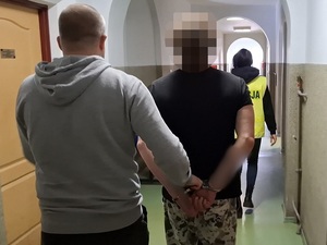 nieumundurowany policjant prowadzi korytarzem mężczyznę, który ma ręce skute za plecami kajdankami. Przed nimi idzie policjantka w kamizelce odblaskowej z napisem "Policja"