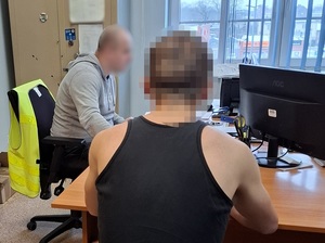 w pokoju przesłuchań na krześle obok biurka policjanta siedzi zatrzymany mężczyzna. Przy biurku przed komputerem siedzi nieumundurowany policjant