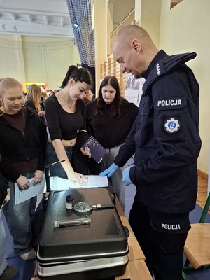 w sali sportowej przy policyjnym stoisku technik kryminalistyki prezentuje zgromadzonej młodzieży sposób zabezpieczania linii papilarnych. Uczennica przykłada dłoń do kartki