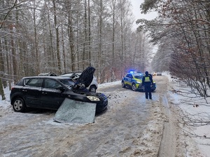 miejsce wypadku drogowego. Pora dzienna. Ośnieżona droga otoczona lasem. W poprzek drogi stoi czarne osobowe auto z licznymi uszkodzeniami. Styka się z nim białe osobowe auto. Na drodze stoi policjant ruchu drogowego. Przed nim znajduje się oznakowany radiowóz