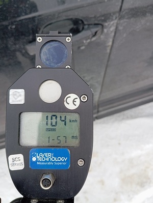 ręczny miernik prędkości z wyświetlonym pomiarem 104 km/h. W tle ciemne auto na zaśnieżonej drodze