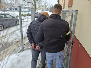 policjant kryminalny prowadzi na zewnątrz budynku komendy mężczyznę, który ma ręce skute za plecami kajdankami