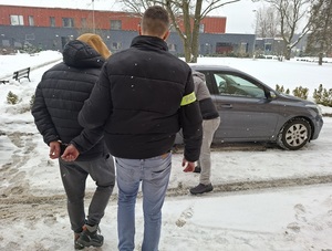 policjant kryminalny prowadzi w stronę nieoznakowanego radiowozu mężczyznę, który ma ręce skute za plecami kajdankami. Drugi policjant otwiera tylne drzwi radiowozu