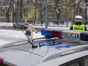 przed szkołą, po drugiej stronie ulicy przy oznakowanym radiowozie stoi policjantka ruchu drogowego, obserwująca ruch na drodze