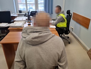 w pokoju przesłuchań na krześle siedzi mężczyzna. Przed nim przy biurku, przed komputerem siedzi policjant