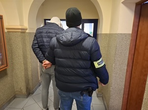 policjant kryminalny prowadzi korytarzem budynku komendy mężczyznę, który ma ręce skute za plecami kajdankami