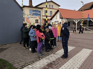 na przejściu dla pieszych stoi dzielnicowa, która rozmawia z dziećmi stojącymi przed przejściem. Obok dzieci stoi dzielnicowy
