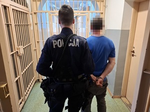 w korytarzu pomieszczenia dla osób zatrzymanych stoi policjant, który trzyma za przedramię zatrzymanego mężczyznę